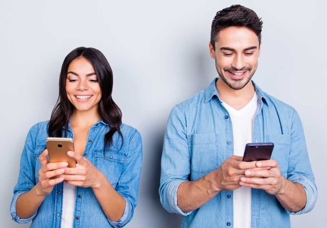 Uma mulher sorridente segurando e olhando para o celular, e ao lado um homem sorridente sorrindo segurando um celular