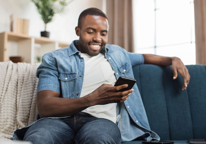Homem sorrindo sentado no sofá enquanto mexe no seu smartphone