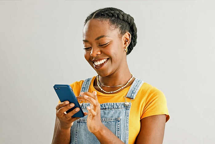 Mercado Pago: mulher negra com transa no cabelo, vestindo blusa amarela, jardineira jeans e acessórios, sorrindo enquanto mexe em seu celular para realizar a liberação do Open Finance na sua conta.