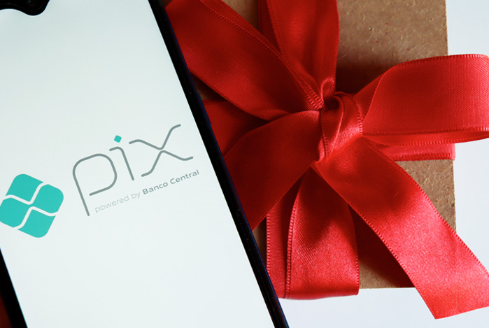 Mercado Pago: celular, com tela aberta no logo do Pix, posicionado em cima de uma caixa de presente, com laço vermelho grande, sugerindo que aquele item foi comprado graças a existência do Pix Parcelado. 