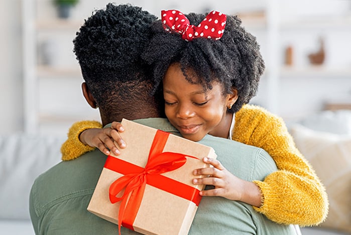Mercado Pago: menina negra com laço vermelho com bolinhas brancas no cabelo, vestindo um suéter amarelo, abraçando o pai, negro, vestindo camiseta verde, enquanto segura com as mãos posicionadas em suas costas, uma caixa de presente de Dias das Crianças com laço vermelho.