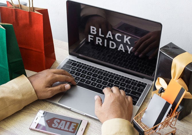 Mercado Pago: Pessoa fazendo compras online na Black Friday. Laptop com a tela mostrando 'Promoções Black Friday', rodeado de produtos e um celular com ofertas.