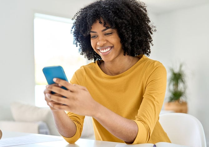 Uma jovem mulher afro-americana feliz e sorridente sentada à mesa em casa, olhando para o celular enquanto observa o seu Pix automático