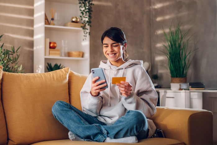Mercado Pago: mulher sentada no sofá, sorrindo, com celular e cartão na mão, verificando seu 13º salário na tela do dispositivo móvel 