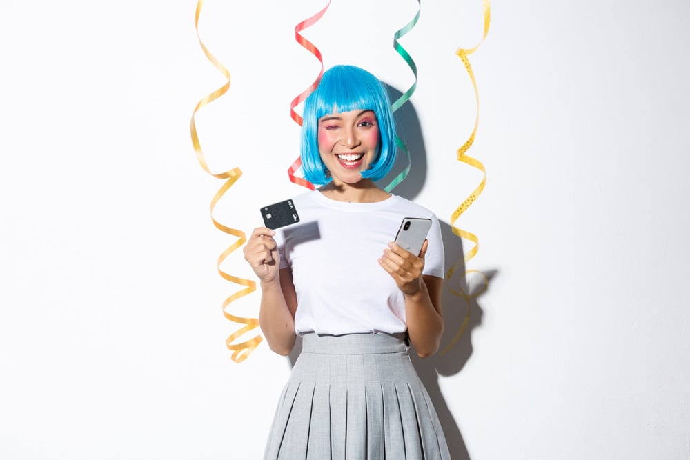 Mulher vestindo camiseta branca e saia cinza rosada, usando uma peruca azul e segurando o celular em uma mão e na outra cartão de crédito Mercado Pago com serpentinas penduradas na parede 
