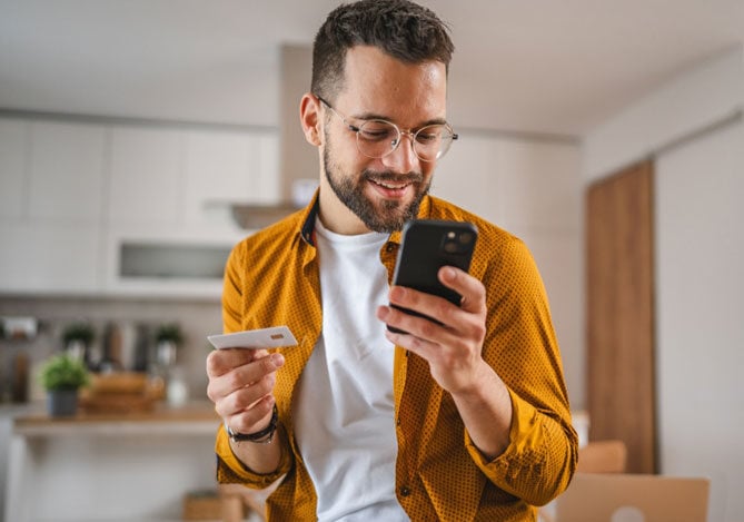 Mercado Pago: homem vestindo camisa amarela e camiseta branca, segurando um celular e um cartão de crédito e realizando compras na Black Friday. Ele está na cozinha de sua casa. 