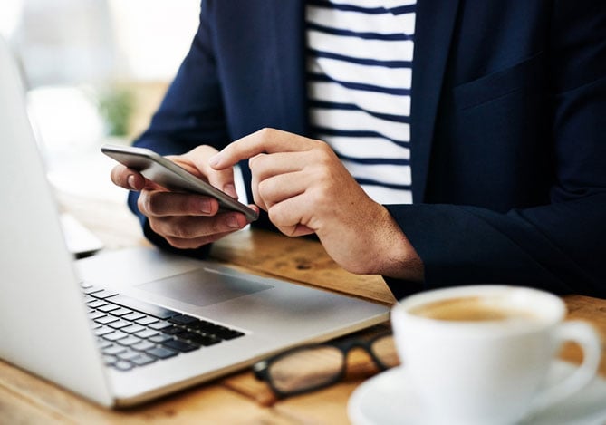 Mercado Pago: mãos de uma pessoa sentada à mesa na frente de um notebook, onde também há um óculos e uma xícara de café. As mãos estão mexendo em um celular para solicitar um cartão virtual.