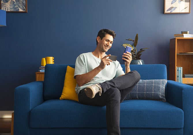 Homem sentado em um sofá azul com almofadas azuis e amarelas. Ele está com as pernas cruzadas, segurando o Cartão Mercado Pago em uma das mãos e na outra o celular para assinar o Meli+ e ter cashback garantido.