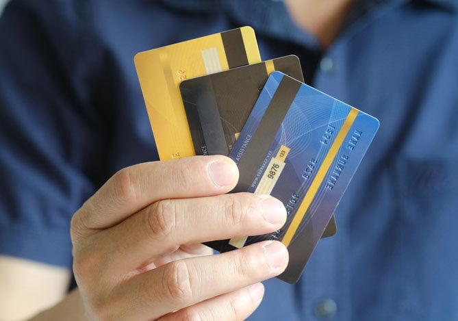 Mercado Pago: imagem de uma pessoa com camisa azul segurando três cartões de crédito nas cores amarelo, preto e azul nas mãos, em que, em um deles, é possível visualizar números que ilustram um código de verificação, ou o CVV do cartão.