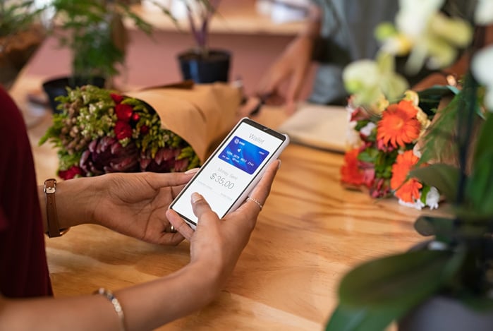 Mercado Pago, mulher sentada à mesa de restaurante com várias flores, com o celular na mão visto de cima, onde está com a carteira digital aberta representando como usar cartão virtual em loja física. 