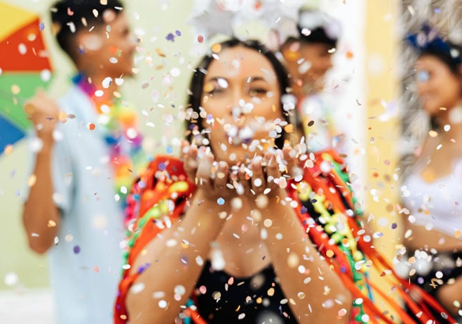 Mulher com adereços de Carnaval soprando confetes enquanto curte a folia sem perder seu controle de orçamento, tudo com a ajuda do Mercado Pago