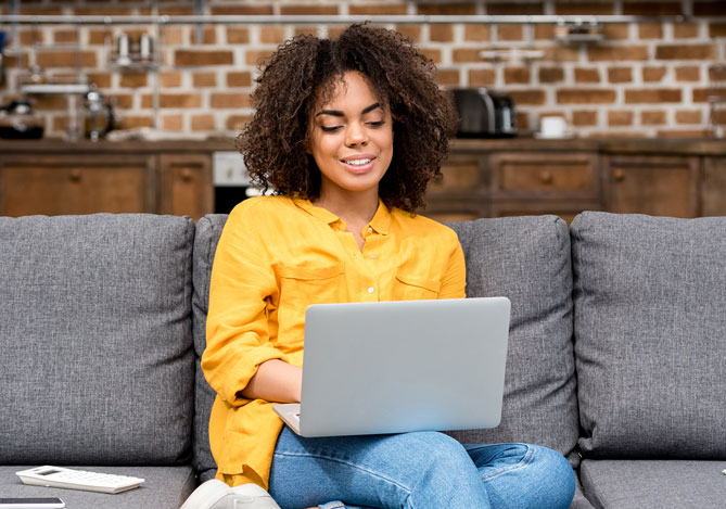 Mercado Pago: mulher sorridente vestindo calça jeans e camisa amarela sentada em um sofá cinza com um laptop aberto em seu colo para verificar sua declaração de imposto de renda do ano passado.