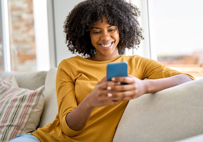Mulher negra com cabelo crespo vestindo blusa de manga amarela sentada no sofá e sorrindo enquanto mexe no celular e verificar como conseguir crédito no banco digital Mercado Pago