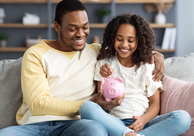 Mercado Pago: Pai e filha abraçados enquanto estão sentados em um sofá colocando moedas em um porquinho sugerindo o processo de educação financeira para se tornar um consumidor consciente.