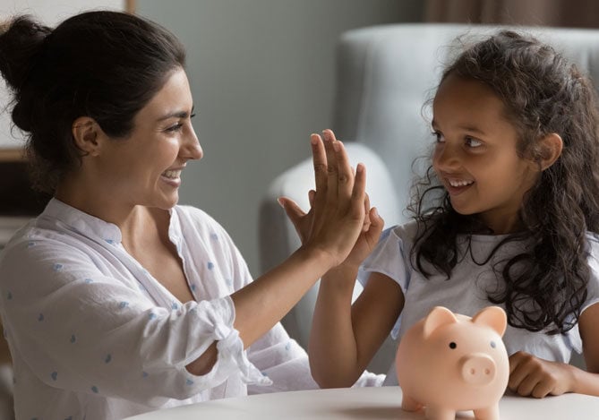 Mercado Pago: Mãe sentada à mesa usando uma blusa branca ensinando educação financeira infantil para a sua filha, que também está com uma camiseta branca. A frente das duas, há um cofrinho de porquinho e elas estão em uma sala de estar. 