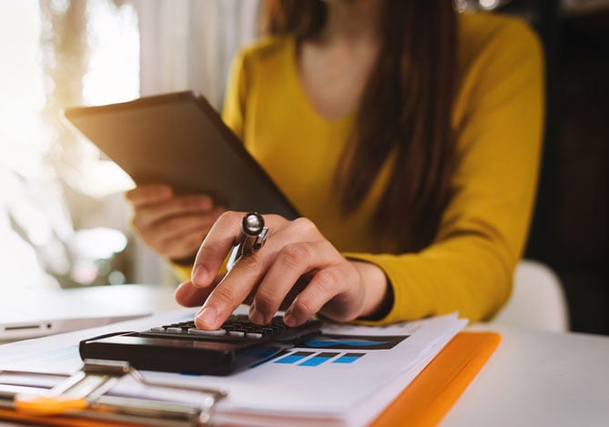 Mulher com blusa amarela conferindo propostas do Empréstimo Mercado Crédito no tablet e fazendo contas na calculadora ao mesmo tempo