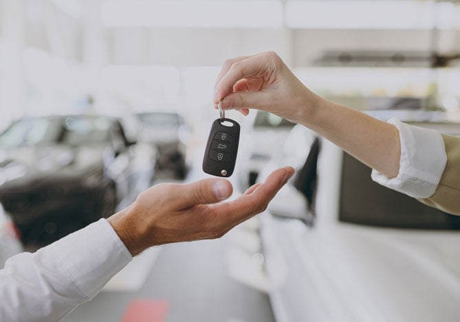 Mercado Pago: imagem da mão de uma pessoa colocando a chave de um carro na mão de outra pessoa. Ao fundo, é possível ver diversos carros, representando uma concessionária de veículos onde é possível financiar um carro.