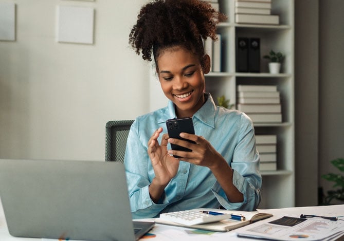 Mercado Pago: imagem de uma mulher vestindo camisa de manga longa azul sentada à mesa de trabalho e sorrindo. Na imagem, é possível ver que a mulher segura um celular em suas mãos para solicitar um empréstimo pessoal por meio de aplicativo. Sobre a mesa há um notebook aberto, algumas folhas, uma calculadora e uma caneta. Ao fundo da imagem, há estantes na parede com livros e pastas de documentos.