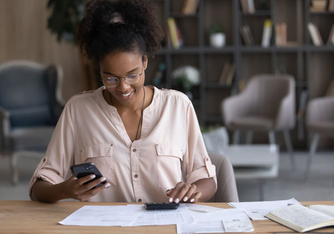 Mercado Pago: Mulher de óculos, com camiseta branca, sentada em mesa com calculadora e papéis apoiados e com celular em sua mão, pesquisando os melhores empréstimos pessoais.