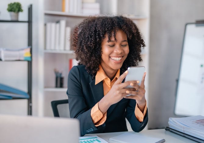 Mercado Pago: imagem de uma mulher sentada à mesa de trabalho utilizando um celular e sorrindo. Na imagem, é possível ver alguns papéis sobre a mesa e uma estante com livros aos fundo, indicando que a mulher está verificando e analisando relatórios das suas finanças pessoais e da gestão do seu dinheiro, feita com a ajuda de inteligência artificial. 