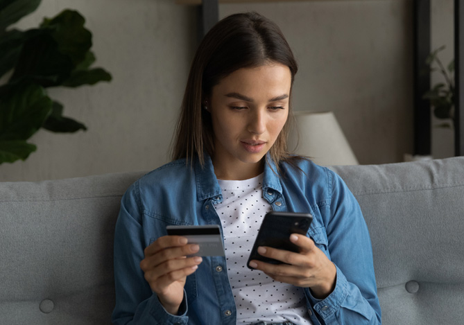 Mercado Pago: mulher sentada em um sofá com o celular em uma mão e um cartão de crédito em outra. Ela olha para a tela do dispositivo móvel de forma séria, pois desconfia que seja alvo de golpes financeiros na internet. 
