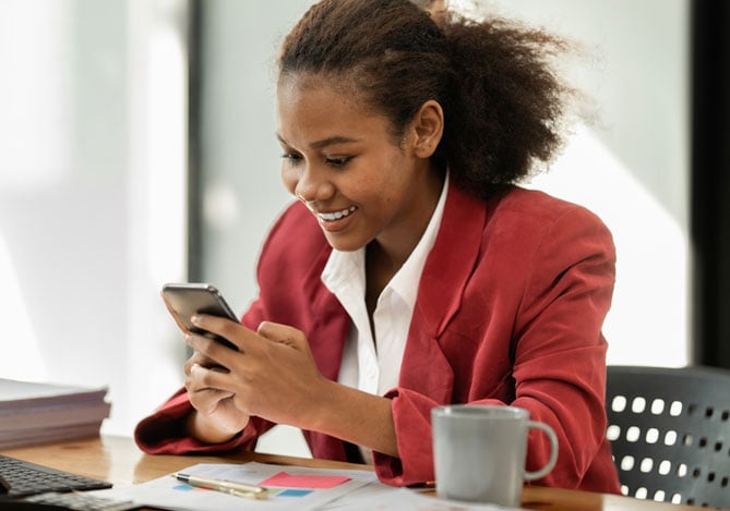 Mercado Pago: mulher sorrindo, sentada à mesa, com um notebook e papéis à sua frente, enquanto ela segura seu celular para realizar um investimento para ganhar dinheiro.