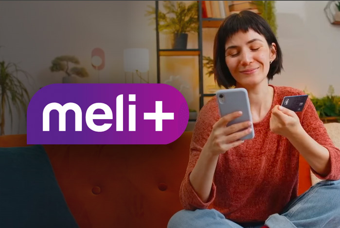 Mulher de cabelo curto, sentada no sofá, vestindo blusa vermelha e calça jeans, segurando nas mãos o cartão Mercado Pago e na outra o celular para avaliar dentre as opções de planos Meli+ qual é o melhor para ela: Essencial, Total ou Mega. 