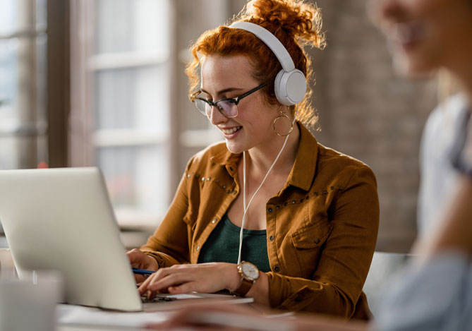 Mercado Pago: imagem de uma mulher vestindo jaqueta marrom e sorrindo sentada à mesa de trabalho digitando em um notebook usando fones de ouvido, enquanto seleciona uma playlist de músicas para ouvir no trabalho e melhorar sua concentração.