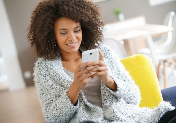 Mercado Pago: imagem de uma mulher vestindo um casaco e com um celular nas mãos. Ela está sentada em uma poltrona amarela na sala de sua casa enquanto consulta em seu celular quais as consequências do não pagamento das parcelas do empréstimo pessoal que realizou para algumas compras pessoais.