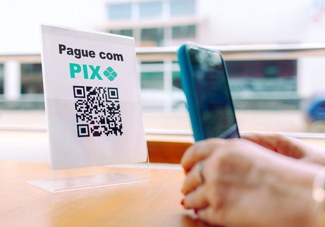 Mercado Pago: imagem das mãos de uma pessoa segurando um celular sobre uma mesa onde há um suporte com o código QR de um comércio para a realização de paramento já dentro das novas regras do Pix. No papel apoiado no suporte, há a frase “Pague com PIX” e abaixo o código QR para pagamento.