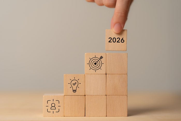 Mercado Pago: dedo de uma pessoa posicionamento blocos de madeira em uma mesa, estampados com ilustrações que sugerem o processo de um planejamento financeiro. Em um dos blocos tem a escrita de “2026”. 