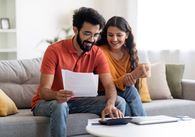 Mercado Pago: um homem e uma mulher, casados, sentados no sofá de casa com uma calculadora e um papel na mão do homem para que façam o planejamento financeiro familiar.