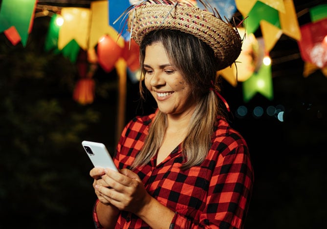 Mercado Pago: mulher em Festa Junina com roupas típicas, como camisa xadrez e chapéu de palha, mexendo no celular.