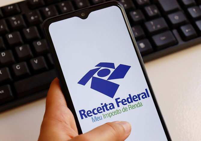 Mercado Pago: imagem da mão de uma pessoa segurando um celular o qual é utilizado para acessar o sistema da Receita Federal e realizar a Declaração do Imposto de Renda. Na tela do celular, é possível ver o logo da Receita Federal, indicando que a pessoa irá acessar o sistema para começar sua declaração.