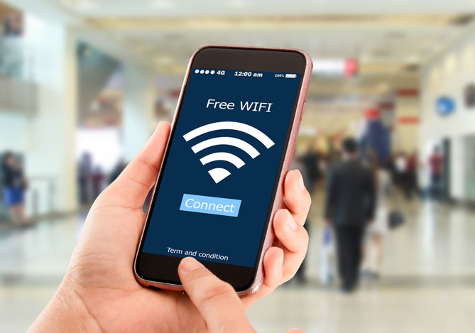 Mercado Pago: imagem de mãos segurando um celular tentando se conectar a uma rede de internet pública. Na tela do celular, é possível ver o símbolo de Wi-fi gratuito e ao fundo, sem foco, é possível ver pessoas em um ambiente público semelhante ao corredor de um shopping.