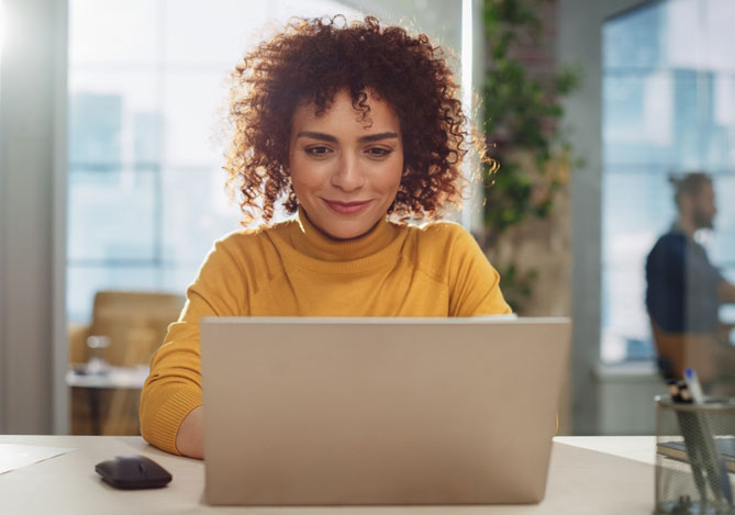 Mulher de cabelo cacheado vermelho vestindo uma blusa amarela e mexendo no laptop para conferir o programa Indique e Ganhe do Mercado Pago para garantir sua renda extra.