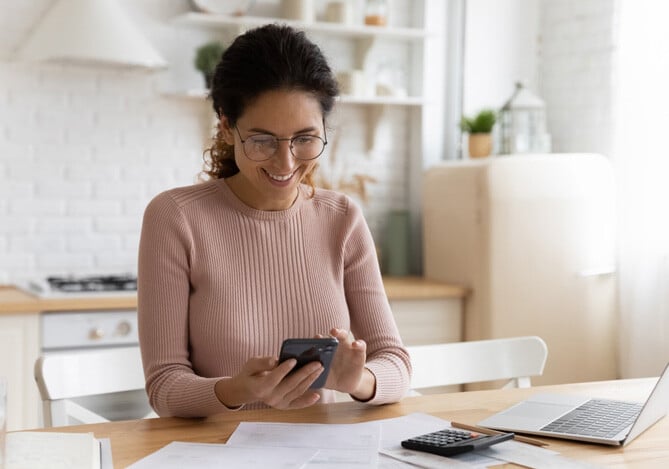 Mulher sorrindo usando óculos de grau, cabelo preso, blusa de lã de manga comprida na cor rosa-claro sentada em uma cadeira branca posicionada na mesa da sua cozinha com contas, calculadora e laptop, mexendo no smartphone para verificar o rendimento CDI da Conta Mercado Pago