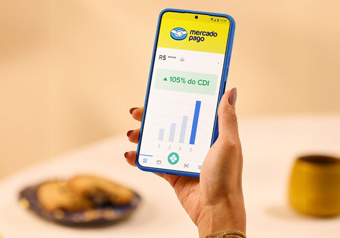 Mercado Pago: mão segurando celular com tela aberta na Conta Mercado Pago mostrando que ela rende 105% do CDI maior que o rendimento da poupança. 