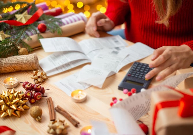 Mercado Pago: imagem das mãos de uma pessoa vestindo uma blusa de manga longa vermelha, sentada à mesa de casa. Sobre a mesa é possível ver alguns enfeites de Natal, caixas de presente e papel de presente, ilustrando que a pessoa está se preparando para o período de Festas. É possível na imagem que a pessoa tem algumas contas impressas e está realizando cálculos com a ajuda de uma calculadora para confirmar se as compras estão dentro do seu planejamento e se sua reserva de emergência, programada para o Natal, será suficiente para cobrir o valor dos gastos.