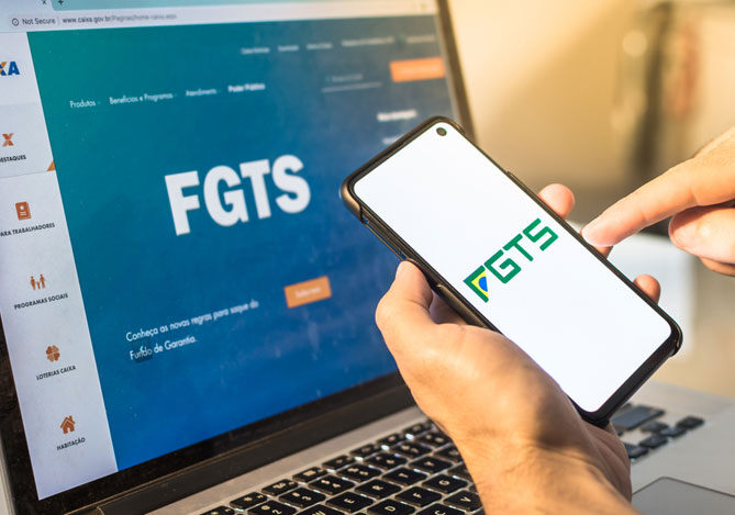 Mercado Pago: imagem da mão de uma pessoa acessando o aplicativo do FGTS para solicitar o Saque-Aniversário em frente a um notebook mostrando também a página do FGTS