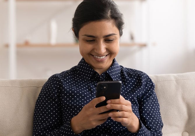 Imagem de uma pessoa sentada em um sofá, segurando um celular nas mãos e sorrindo, enquanto organiza suas finanças para destinar uma parte da segunda parcela do décimo terceiro para o pagamento de contas e guardar outra parte no Reservas do Mercado Pago e deixar esse valor rendendo ao longo do ano.