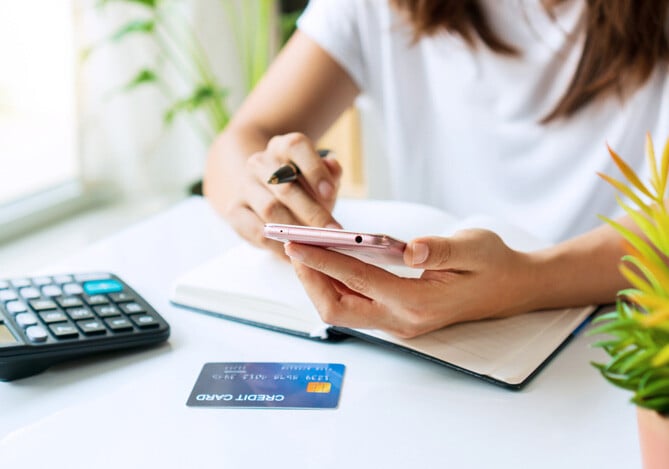 Mercado Pago: Mulher sentada à mesa vestindo uma blusa branca e segurando um celular, pesquisando sobre as taxas e juros do seu cartão de crédito. Na mesa, há um caderno, um cartão e uma calculadora. 