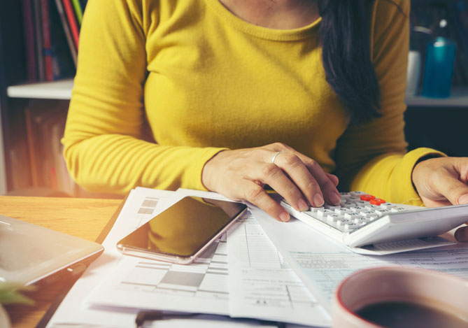 Mãos de uma mulher vestindo uma blusa amarela do Mercado Livre, parceiro do banco digital Mercado Pago, mexendo em uma calculadora para fazer cálculos relacionados aos tipos de impostos em investimentos