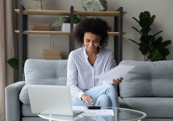 Mercado Pago: imagem de uma mulher sentada no sofá de sua casa em frente a uma mesa de centro sobre a qual há um notebook aberto, algumas folhas e uma calculadora. Em uma das mãos, a mulher segura alguns papéis e com a outra ela digita na calculadora, enquanto escolhe entre os tipos de orçamento que está pesquisando com seu notebook para verificar qual melhor se adapta à sua realidade financeira.