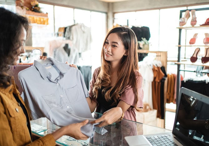 Mercado Pago: imagem de uma mulher em uma loja de roupas e sapatos segurando uma camisa polo no caixa junto com a funcionária da loja para realizar uma troca de presentes de natal. Ao fundo da imagem, é possível ver outras peças de roupa e sapatos nas gôndolas.