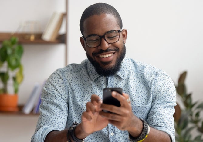 Mercado Pago: homem usando óculos e camisa azul estampada, sorrindo enquanto mexe no celular para usar o seu banco digital para investir.