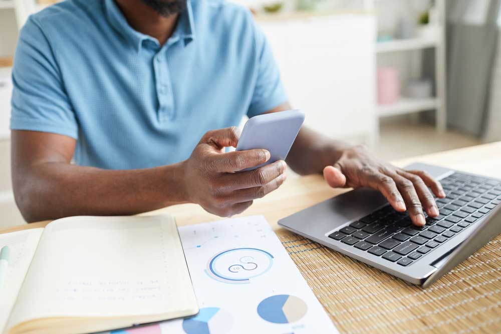 Homem com um celular na mão e digitando no computador na outra